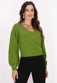 Grüner strukturierter Pullover mit V-Ausschnitt und Ballonärmeln, der einen taillierten Bund hat. Kombiniert mit schwarzen Hosen.