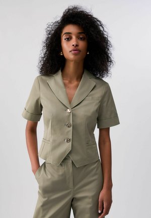 Donna con capelli ricci che indossa un blazer kaki su misura a maniche corte con tre bottoni e pantaloni abbinati a vita alta, in piedi con una mano in tasca.