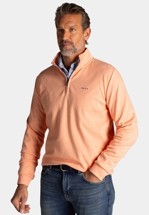 Man with short gray beard wearing peach quarter-zip pullover over blue collared shirt and blue jeans, standing with one hand in pocket.