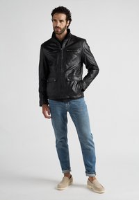 Schwarze Lederjacke mit hochgeschlossenem Kragen, Reißverschlusstaschen und zwei vorderen Klappen. Getragen mit blauen Jeans und hellen Slippern.