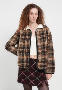 Καρό μπουφάν σε καφέ και μαύρο με κρεμ γιακά, μπροστινά κουμπιά και πλαϊνές τσέπες, σε συνδυασμό με μπορντό φούστα με σχέδιο αργκλάι. Υφασμα από υφή μαλλιού.