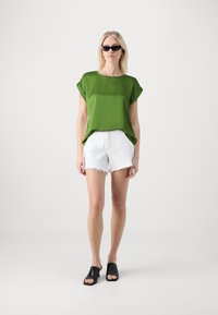 Grønn kortermet topp laget av glatt stoff, kombinert med hvite shorts med frynset kant og svarte åpen-tå mules, som skaper et uformelt utseende.