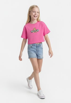 Ragazza sorridente che cammina, indossa una maglietta rosa corta con grafica dell'orso e shorts di jeans, scarpe da ginnastica bianche, su uno sfondo bianco semplice.