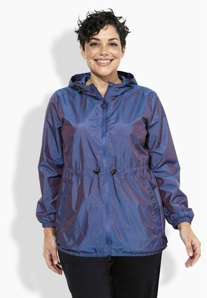 Uśmiechnięta osoba z krótkimi kręconymi włosami, ubrana w niebieską wiatroodporną kurtkę z kapturem zapinaną na zamek, z regulacją w talii, oraz czarne spodnie.