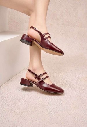 Femme portant des chaussures slingbacks à talon bas carré de couleur rouge foncé avec des brides à double boucle, debout sur un sol clair texturé près de marches blanches.