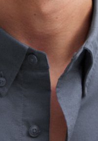 Camicia grigia con colletto a punta, bottoni visibili e un tessuto liscio e leggermente strutturato. Inquadratura ravvicinata sulla zona del colletto.
