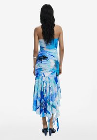 Robe bleue sans bretelles avec un ourlet superposé et volants, ornée d'un motif floral aquarelle dans des nuances de bleu variées. Portée avec des talons bleus.