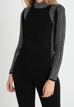 Haut noir à manches longues avec col montant orné de clous argentés densément disposés sur les manches, les épaules et le col, porté avec un pantalon noir.