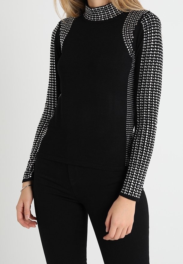 Haut noir à manches longues avec col montant orné de clous argentés densément disposés sur les manches, les épaules et le col, porté avec un pantalon noir.