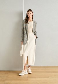 Geruite blazer in grijs en zwart, beige aansluitend top, roomkleurige hoge taille rok met een split aan de voorkant, en lichtgekleurde sneakers; beige tas.