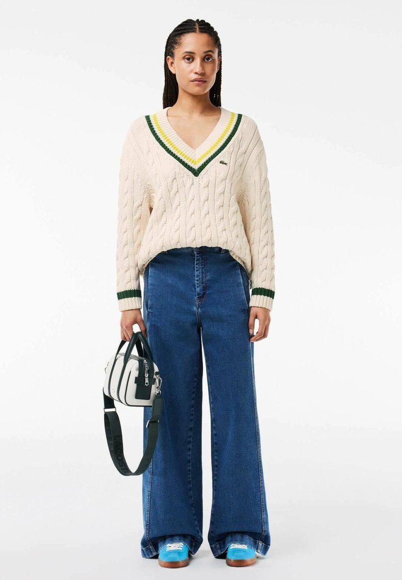 Pull en câble crème à col en V vert et jaune, associé à un jean bleu à jambes larges. Tient un sac blanc aux accents vert foncé.