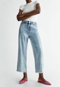 Vaaleansiniset raidalliset farkut, leveälahkeiset, joissa on revitty helma, yhdistettynä vaaleanpunaisiin litteisiin sandaaleihin, joissa on kudottu muotoilu. Lyhythihainen valkoinen ribbipaita.