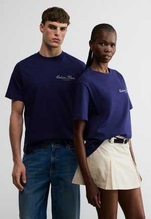 Δύο μοντέλα φοράνε μπλε ναυτικό t-shirts Calvin Klein, το ένα με μπλε τζιν και το άλλο με μπεζ πλισέ φούστα και μαύρη ζώνη.