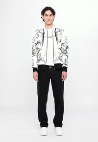Giacca con cappuccio con motivi floreali neri su fondo bianco, polsini neri, abbinata a una maglietta bianca e pantaloni neri, indossata con sneaker bianche.