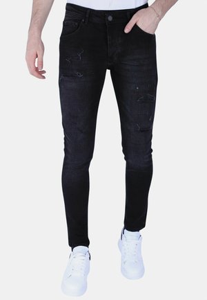 Man draagt zwart gescheurde skinny jeans en witte sneakers, met één hand in de zak en een wit overhemd ingestopt.