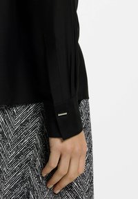 Blusa nera con dettagli ai polsini in un'accento metallico rettangolare, abbinata a una gonna con un motivo a spina di pesce in bianco e nero.