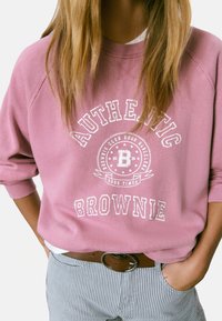Persona que lleva un sudadera rosa con el texto "Authentic Brownie", pantalones de rayas azules y un cinturón marrón, con las manos en los bolsillos y el cabello largo y rubio visible.