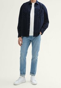 Camicia a righe navy con bottoni e due taschini sul petto, abbinata a jeans azzurri chiari e sneakers bianche. Vestibilità casual con un design rilassato.