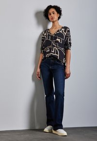 Blusa nera e beige a fantasia con scollo a V, maniche lunghe, abbinata a jeans svasati blu scuro e sneakers bianche.