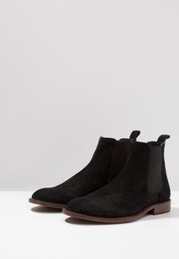 Bottes Chelsea en daim noir avec panneaux latéraux élastiques, bout arrondi et semelle en cuir marron. Équipées de languettes pour un enfilage facile.