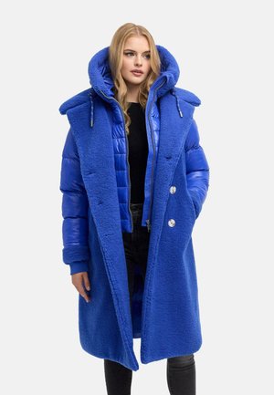 Young woman wearing a long blue padded and fleece winter coat with a large hood, black top, and black pants, standing and looking right.