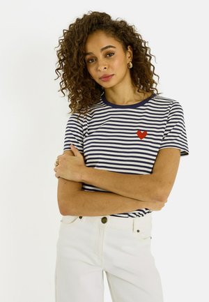 Joven con cabello rizado, vestida con una camiseta de rayas azul marino y blanca con un emblema de corazón rojo y pantalones blancos, con los brazos cruzados.