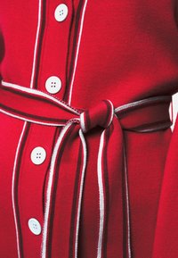 Czerwona tkanina odzieżowa z białymi paskami, z zapięciem na guziki i związanym paskiem. Godne uwagi detale to cztery białe guziki i teksturowany dzianinowy materiał.