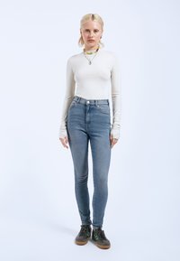 Top de manga larga gris claro combinado con jeans skinny de cintura alta azul claro. El modelo lleva zapatos oscuros con acentos verdes. Diseño minimalista.