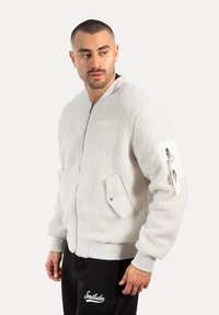 Hombre vestido con chaqueta de forro polar blanca con cremallera y bolsillos, y pantalones negros, mirando hacia su izquierda contra un fondo liso.