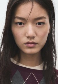 Prim-plan al unei persoane cu păr lung și închis la culoare, purtând un pulover bordo cu un model geometric în gri și alb. Piele netedă, expresie neutră.