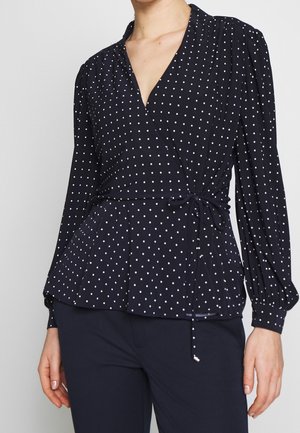Femme portant un chemisier cache-cœur bleu marine à pois blancs et manches longues, associé à un pantalon bleu marine foncé.