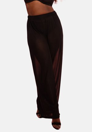 Woman wearing black sheer wide-leg pants and black open-toe heels, posing against a plain light background.