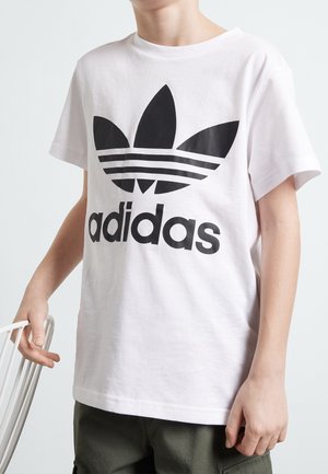 Enfant portant un t-shirt Adidas blanc avec un logo noir, debout et tenant le dossier d'une chaise blanche.