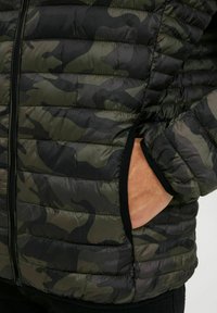 Veste matelassée camouflage avec un design en vert foncé et noir, dotée d'une fermeture éclair à l'avant et de poches latérales. Tissu texturé.