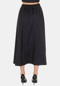 Gonna nera di media lunghezza con righe verticali bianche, vita elasticata, tessuto fluente e texture liscia. Indossata con scarpe nere con tacco alto.