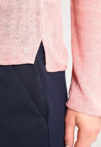 Vaaleanpunainen neulepaita, jossa on teksturoitu pinta, sivuväli ja hopeinen logoakseli. Yhdistetty tumman laivastonsinisen kankaan kanssa.