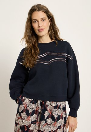 Frau mit langen welligen Haaren, die einen marineblauen Pullover mit gestreiftem Zickzackmuster und einen floralen Rock trägt, steht mit einer Hand in der Tasche.