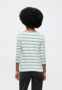 Γυναίκα με αφρο hairstyle, φορώντας λευκό και πράσινο ριγέ μακρυμάνικο μπλουζάκι και μαύρα τζιν, στραμμένη μακριά σε ένα απλό φόντο.