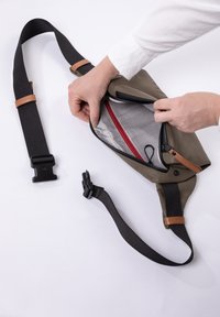 Olijfgroene schoudertas met een zwarte strap en bruine leren accenten, voorzien van een met rits sluitende zak met rode voering en een gaasverdeling binnenin.