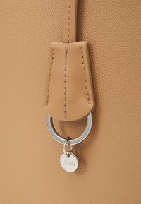 Détail d'un sac en cuir texturé couleur fauve avec languette cousue et anneau argenté tenant un petit pendentif rond gravé "LIU JO."