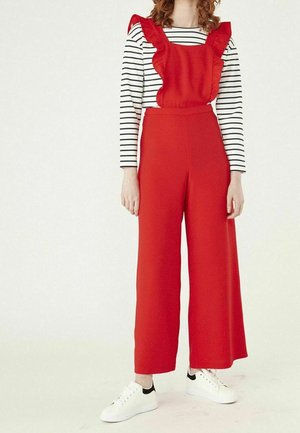 Femme portant une salopette rouge à jambes larges avec des bretelles volantées et un haut à manches longues noir et blanc rayé, associée à des baskets blanches.