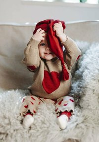 Maglione tan con un grande cuore rosso, leggings rossi a fantasia e scarpe bianche. Bambino tiene un cappello rosso con un pon pon, seduto su una coperta soffice.