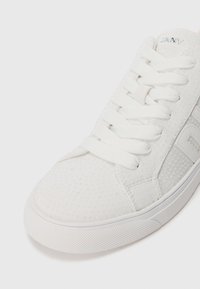 Sapatilhas brancas com textura perfurada, cordões planos e solas de borracha. A lateral exibe a marcação em cinza claro. Design limpo e minimalista.