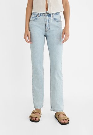 Femme portant un jean droit taille haute bleu clair, un top court blanc et des sandales ouvertes beiges avec deux boucles sur fond blanc.