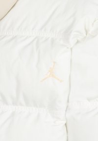 White puffer jacket featuring a textured finish, elasticated waist, and an embossed beige Nike Air Jordan logo on the lower left.