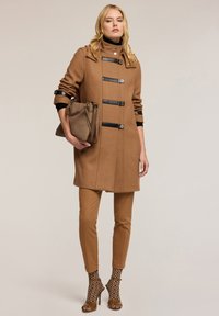 Abrigo de lana marrón con acentos negros, botones y un cuello alto. Combinado con pantalones beige y calcetines negros con estampado, sosteniendo un bolso marrón.