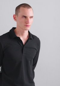 Μαύρο πουκάμισο πόλο με μακριά μανίκια, γιακά και κουμπότρυπα. Λείος υφασμάτινος σχεδιασμός, εφαρμοστός, με ελάχιστες λεπτομέρειες. Ανοιχτό φόντο.