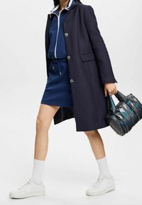 Cappotto blu con bottoni, abito sportivo blu sotto, sneakers bianche e una borsa nera lucida e multicolore.