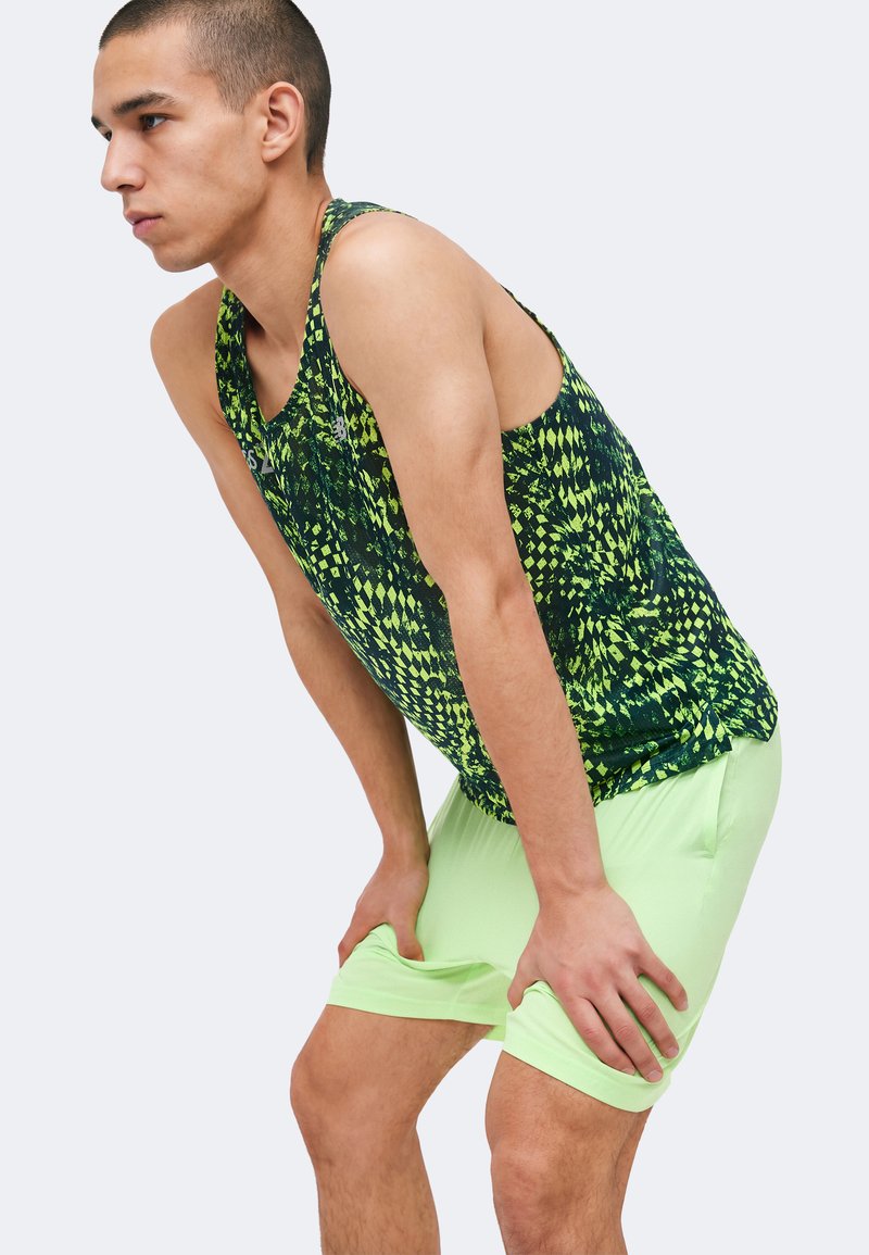 Jeune homme penché en avant, les mains sur les genoux, portant un short vert néon et un débardeur sportif sans manches à motifs.