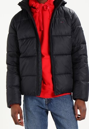 Uomo che indossa una giacca imbottita nera trapuntata sopra una felpa con cappuccio rossa con coulisse e jeans blu, in piedi con le braccia rilassate lungo i fianchi.